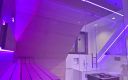 Glassauna mit Eckverglasung und Dachschräge, Innenverkleidung aus waagerechten Saunapaneelen in Hemlock, Einrichtung in Thermo-Espe mit LED-Lehnenbeleuchtung - Seitenansicht mit schwebender Saunabank, LED-Beleuchtung violett