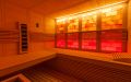Massivholz-Sauna aus Fichte mit Eckeinstieg, Ganzglaselement, Infrarotstrahler und beleuchteter Salzsteinwand - Einrichtung und gelb-rot beleuchtete Rückwand