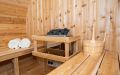 Fass-Sauna aus weißem Zedernholz mit kleiner Veranda - Innenansicht mit Saunaofen, Saunabänken, Liegetüchern und Aufgusseimer