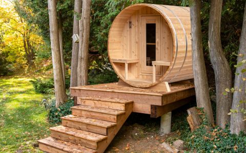 Fass-Sauna aus weißem Zedernholz mit kleiner Veranda - Außenansicht von schräg rechts, platziert auf einem Holzpodest im Wald